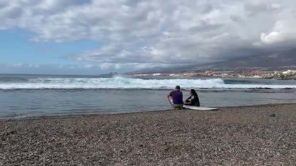 サーファーのカップルは海に座って泳ぎについて話し合います サーフィンの後に話す海の背景に女の子を持つ男 — ストック動画