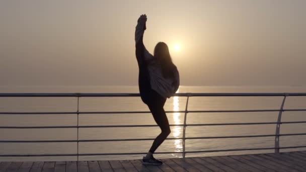 Девушка демонстрирует невероятную гибкость тела. Она растягивает ноги, стоя на набережной возле моря. Утреннее солнце. Сгибается в арку, стоящую в вертикальной бечевке — стоковое видео
