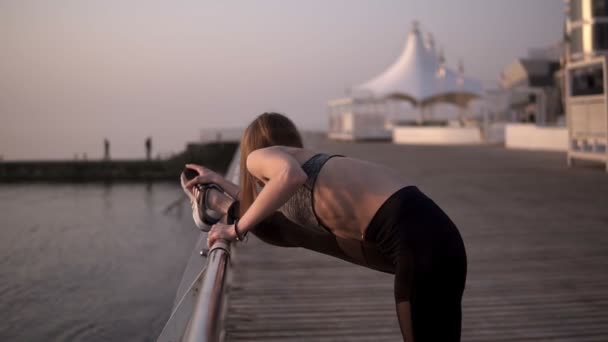 Tunna och passar unga kvinnliga i sportkläder sträcker benmusklerna på ett havsområde. Klädd i svarta leggings och grå BH. Lutar sig på en tvärslå. Sidovy — Stockvideo