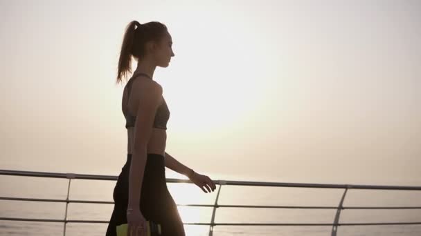 Gorgeous side view of a pretty girl walking on the road along the sea, the sun shines quietly. A girl in black leggings and top. Pony tail. Carefree — Stock Video