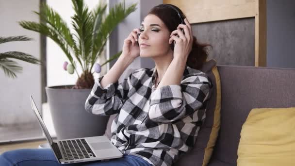 Linda, jovem mulher relaxa sentado no sofá, coloca fones de ouvido e liga a música no laptop. Sorri e move-se ao ritmo da música. Movimento lento — Vídeo de Stock