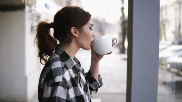 Hermosas chicas vista de pie cerca de las ventanas. Beber un té con una taza blanca. Vista lateral. Fondo borroso — Vídeo de stock