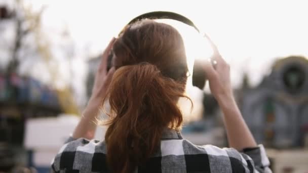 Imágenes de la parte trasera de una chica con una cola de pelo castaño de pie una camisa a cuadros oover contra el sol brillante. Ponerse los auriculares — Vídeos de Stock