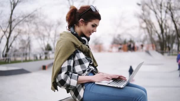 Een jonge trendy brunette is zittend op een bankje met een laptop op haar knieën, te typen. Koptelefoon op de nek, glazen. In het park zijn kinderen schaatsen op scooters en fietsen. Zijaanzicht — Stockvideo