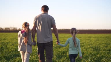 İki küçük kızı ile yeşil çayır tarafından yürüyüş bir güçlü, Genç Baba arka görünümü. Onları her iki eliyle tutarak. Bahar günü. Görüntüleri izleme