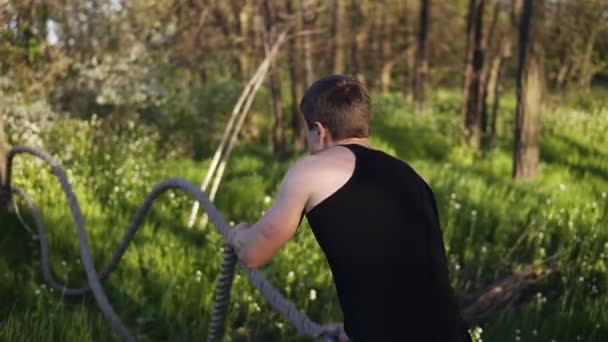 L'uomo dello sport si allena con le corde ondulate. Un duro lavoro. T-shirt nera. Parco verde con alberi. Giornata di sole. Vista posteriore — Video Stock