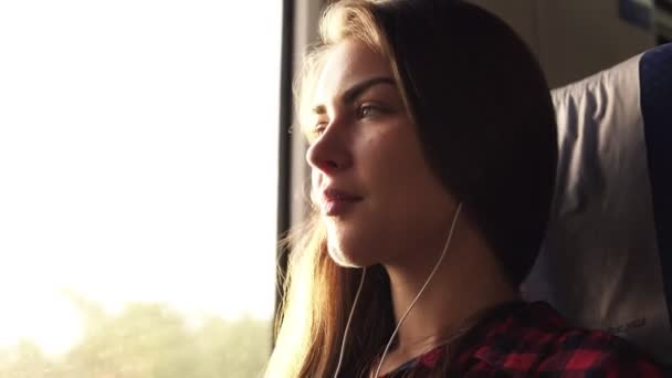 Vista lateral de una hermosa chica caucásica que va a algún lugar en tren. Escucha música en los auriculares. Mujer pensativa. Sonriendo. El sol brilla — Vídeos de Stock