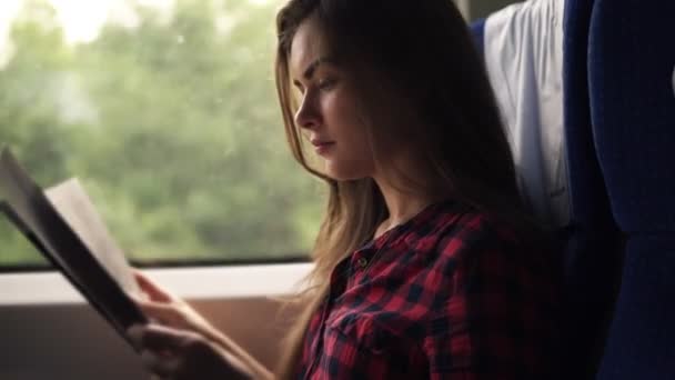 Una chica joven y bonita viajando en tren moderno. Sentada junto a la ventana y leyendo en camisa a cuadros. Vista lateral. Ventana borrosa en movimiento — Vídeos de Stock