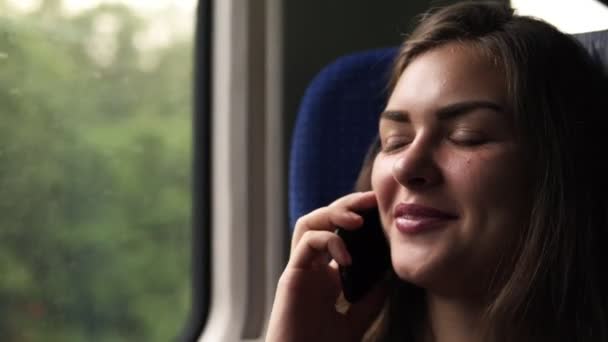 Zbliżenie twarzy womans. Ładna dziewczyna jest podróż pociągiem. Rozmowy na jej telefon komórkowy. Uśmiechnięty, zrelaksowany. Ruchomy obraz natury w oknie — Wideo stockowe