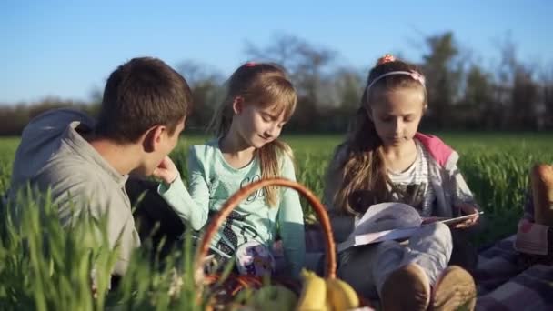 Прекрасный, ветреный день. Отец с двумя дочерьми устраивает пикник на зеленом поле. Маленькая девочка кормит своего отца печеньем. Закрыть — стоковое видео