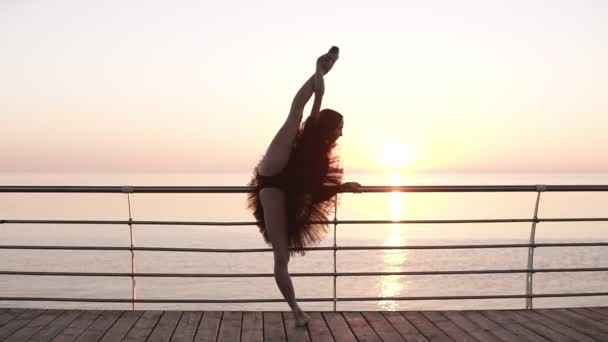 Eine Ballerina steht neben dem Geländer an der Promenade. Sie dehnt sich und beugt ihre Beine in einem senkrechten Faden. Sonne scheint. schöner Morgen Meerblick — Stockvideo