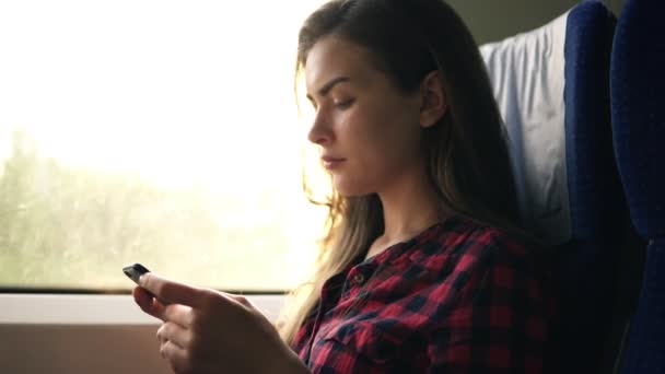 Aantrekkelijke vrouw in plaid shirt gaat met de trein, zitten naast het venster en serieus op zoek op haar mobiele telefoon. Zijaanzicht. Vervoer, beweging — Stockvideo