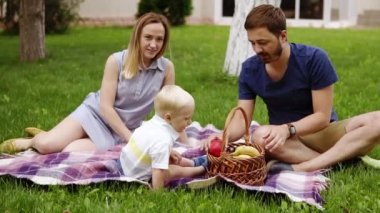Güneşli bir günde bir ekose piknik yeşil güzel çayır üzerinde oturan mutlu bir aile. Oğulları yakınında oturuyor. Pikniğin tadını çıkarın. Açık havada