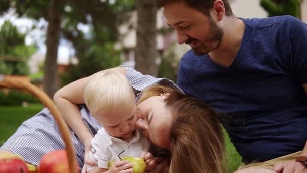 Närbild av en unga föräldrar som sitter nära varandra med deras lille son. Picknick ute på en pläd. Picknick-korg med äpplen. Leende och lycklig familj — Stockvideo