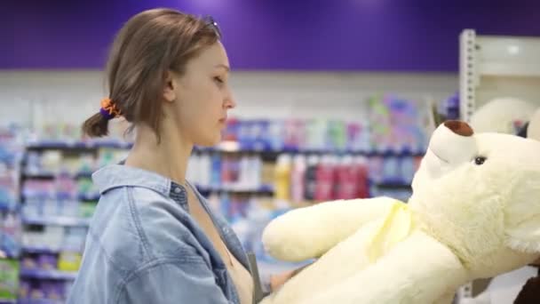 Imágenes de cerca de la chica en busca de los juguetes blandos en el estante en el supermercado. Ella está decidida a elegir. Estante con surtido de juguetes de peluche. Chica con ropa casual. Vista lateral — Vídeos de Stock