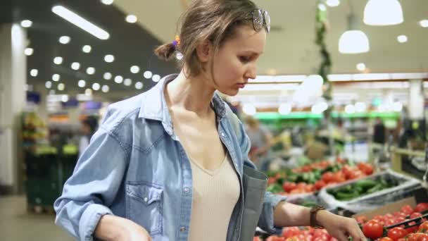 Flickan, hemmafru shopping på stormarknaden. Väljer färska tomater på en gren, sätter dem i en cellofan påse, binder dem och sätter dem i en vagn. Fulla människor lagra. Handhållen — Stockvideo