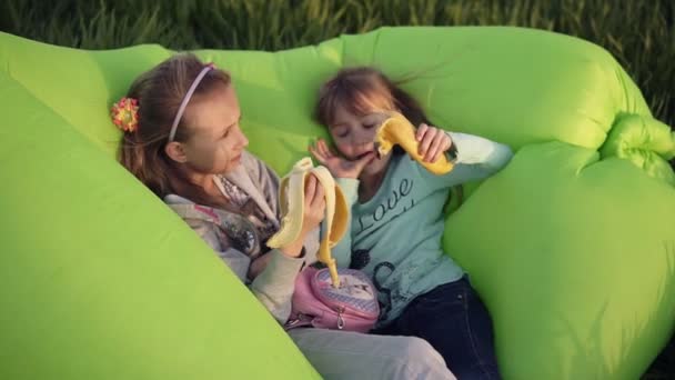 Két kis nővére feküdt felfújható zöld kanapén szabadban. Pihenési, étkezési banán. Kis egy nyalás ujjak. Ízletes. Süt a nap. Zöld fű — Stock videók