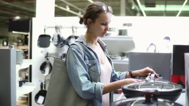 Filmagem portátil de uma jovem mulher em compras de camisa jeans. À procura de coisas da cozinha. Examinando vários potes e panelas com tampo de vidro. Supermercado, loja. Vista lateral — Vídeo de Stock