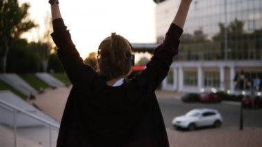 Şık bir kız açık havada modern arka plan üzerinde bina ile sokakta dans görüntüleri. Müziğin, ellerini yükselterek kadın komik bir biçimde hareket. Mutlu, gülümseyen. Dusk