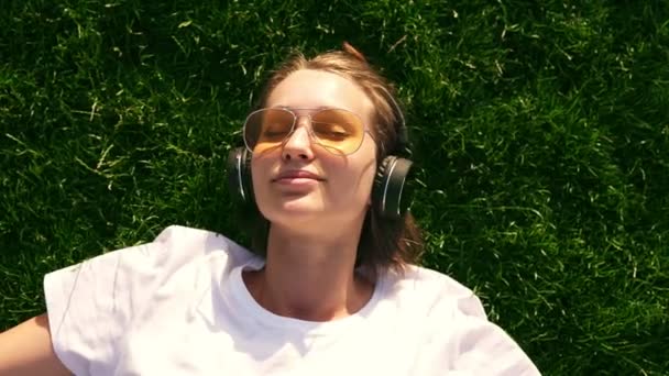 女孩听音乐流着耳机在夏天在草地上。绿草。微笑的女孩在黄色的太阳镜和白色 t恤衫。从顶部拍摄的画面 — 图库视频影像
