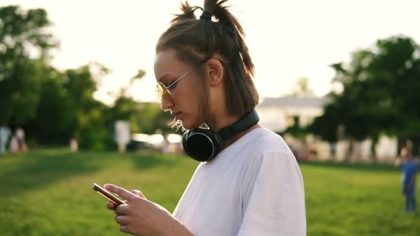 Vista lateral de uma menina fantasia em camiseta branca digitando em seu telefone celular. Fones de ouvido pretos no pescoço. Passar tempo no parque verde. Sol brilha no fundo — Vídeo de Stock