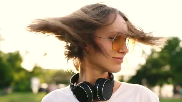 Jonge mooie meisje met kort eerlijke haar schudt haar hoofd geïsoleerd over groene park achtergrond. Kaukasische vrouw in gele zonnebril en zwarte wareless koptelefoon op haar nek. Vooraanzicht, slow-motion — Stockvideo