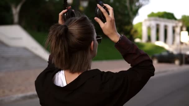 Vista trasera de una joven chica moderna en negro casual caminando por el parque o la calle. Mirando por encima de su hombro y sonriendo. Se pone audífonos desprevenidos. Tiempo libre — Vídeos de Stock