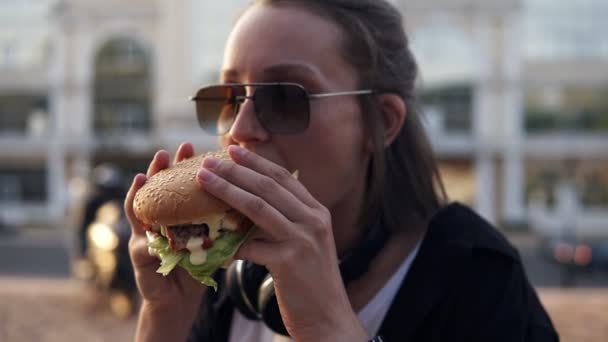 Attraktiv ung kvinna, ler glatt, innehar välsmakande hamburgare i två händer och äter. Klädd i casual outfit, i solglasögon. Utomhus, nära spegeln byggnaden. Närbild — Stockvideo