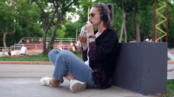 Κομψή, νεαρή κοπέλα βρίσκεται σε ένα πάρκο στο πάτωμα με τα πόδια σταυρωμένα. Πίνοντας ένα αναψυκτικό. Μια γυναίκα με γυαλιά ηλίου και ακουστικά ακούει μουσική, απολαμβάνοντας χρόνο. Αργή κίνηση — Αρχείο Βίντεο