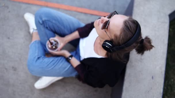 Mujer joven relajada sentada en el suelo al aire libre y escuchando la música. Vestido con jeans azules, camiseta blanca y abrigo negro. Sostiene vidrio transparente en sus manos. Filmación desde arriba — Vídeos de Stock