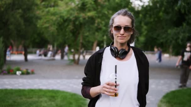 Cheerful, smiling young woman walking outdoors. Casual style. Fashion girl in dunglasses and black headphones on neck. Wind blowing on her face. Drinking a soft drink. Green park. Front view — Stock Video