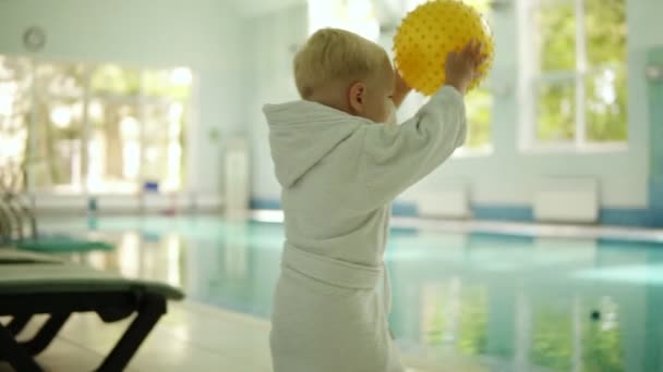 Netter kleiner Junge wirft einen gelben Ball in den Pool des Freizeitzentrums. Schwimmbad drinnen. Junge trägt kleinen weißen Frottee-Bademantel. Zeitlupe — Stockvideo