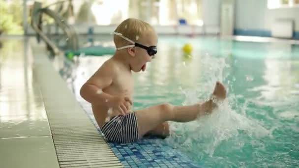 A small blond boy sits on the edge of the pool in swimming trunks and glasses and sprinkles water with his feet. Splashing the water. Having fun. Indoors — Stock Video