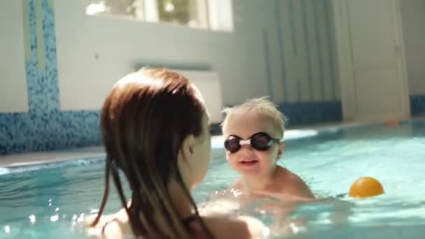 Junge Frau, die ihren Sohn hält und ihm das Schwimmen beibringt. Stützen über dem Wasser. Hallenbad. Kleiner Junge mit Schwimmbrille — Stockvideo