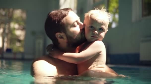 Retrato de un padre y un hijo pasando tiempo juntos en la piscina. Los rayos del sol sobre ellos. Padre está abrazando a su hijo, abrazándolo, besándolo. El niño está en manos de los padres. Piscina cubierta — Vídeo de stock