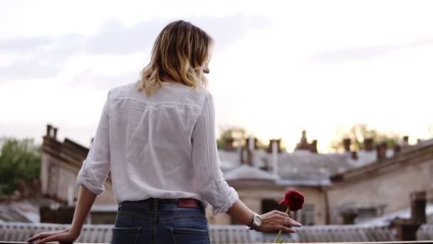 Sensuell, slank kvinna i jeans och vit skjorta står på balkong med röd blomma i handen. Blond tjej. Vacker stad perspektiv. Baksidan — Stockvideo