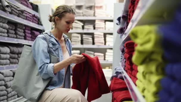 Femme caucasienne choisir des serviettes de grande variété dans la rangée. Les étagères en magasin sont remplies de différentes serviettes de toutes les couleurs. Fille déplie une grande serviette rouge de l'étagère inférieure — Video