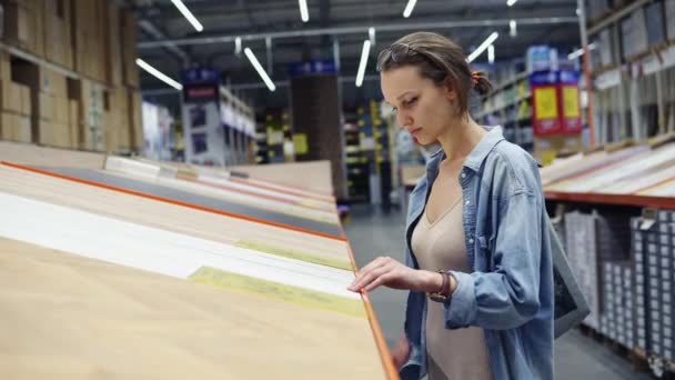 Uma jovem caucasiana em um grande hipermercado, no departamento de materiais de construção e madeira. Tocando as superfícies de madeira, laminado. Concentrado escolhe o produto. Vista lateral — Vídeo de Stock