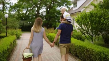 Genç aile gündüz otdoors yürüme. Evler bölgesi. Piknik sepeti üzerinde elini tutan anne. Babam yakaladığında onun omuzlarında bir evlat. Arka görünüm