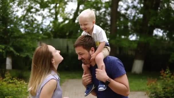 Feliz alegre joven familia padre, madre e hijo pequeño que se divierten al aire libre, jugando juntos en el parque de verano, campo. Mamá, papá e hijo sobre los hombros de los padres, disfrutando de la naturaleza afuera. Movimiento lento — Vídeo de stock