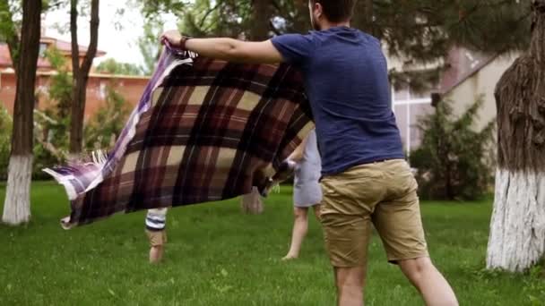 Padre en pantalones cortos marrones y camisa azul está extendiendo la tela a cuadros para el picnic. Feliz madre está jugando con su pequeño hijo en el fondo borroso. Hierba verde. Movimiento lento — Vídeo de stock