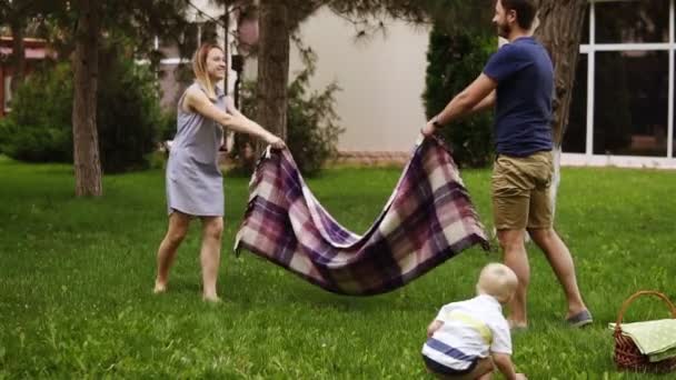 Movimento lento de uma jovem família com um filho num relvado verde. Os pais estão a espalhar o xadrez. Emoção. Menino loiro em uma perspectiva de frente — Vídeo de Stock