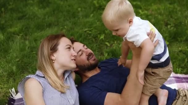 3 つの美しい家族のハイアングル映像。彼の幼い息子と遊んで、緑の草の上に横たわっています。幸せな生活。晴れた日 — ストック動画