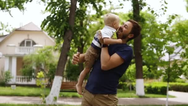 Giovane padre con la barba in abiti casual sta tenendo in braccio il suo piccolo figlio biondo. Ragazzo a piedi nudi. Un uomo che bacia suo figlio. Cottage Street sullo sfondo. Verde e luminoso — Video Stock