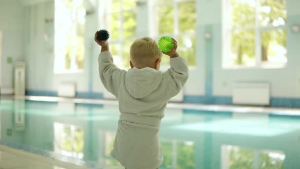 Προβολή πίσω από ένα μικρό αγόρι στο μπουρνούζι ρίχνουν μπάλες από τα δύο χέρια στην πισίνα. Παιχνιδιάρικο. Έξω από την πισίνα. Καθαρό νερό — Αρχείο Βίντεο