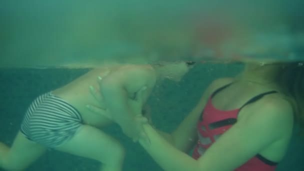 Vista lateral, una inyección submarina de mujer joven en traje de baño rosa que apoya a su hijo en la piscina de agua. Enseñando a nadar. Piscina cubierta — Vídeo de stock