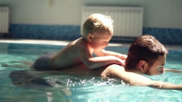 Mladý muž veslování pod vodou se synem blondýna na zadní straně. Malý chlapec je nad vodou. Vnitřní plavecký bazén. Zpomalený pohyb