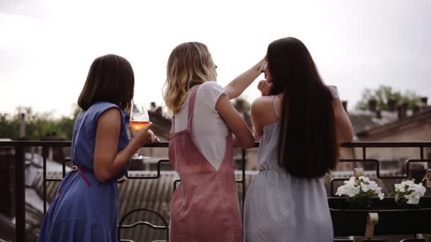 Три подруги наслаждаются девичником на террасе, вид сзади. Модная, современная одежда. Красивый вид на город на фоне — стоковое видео