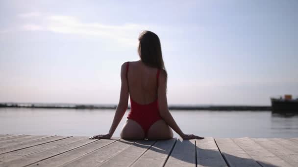 Jolie brune mince sexy étonnante assise sur une jetée en maillot de bain rouge, images du dos, corps mince parfait, cheveux longs. Style bikini. Bord de mer — Video