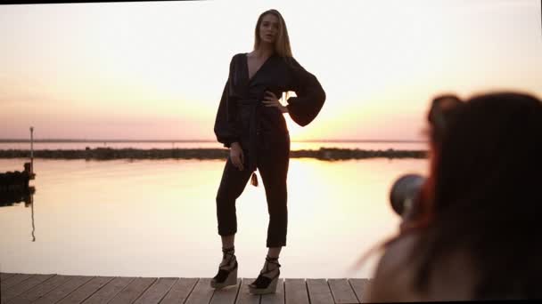 Modelo alto bonito posando para a câmera em rompers pretos e sandálias de pé em um comprimento total na frente do mar. Conceito de fotofotografia — Vídeo de Stock